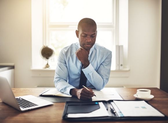 Man Looking At Paperwork On A Table With A Laptop Nearby