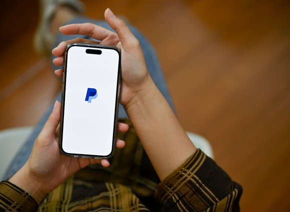 Person Holding A Smart Phone With The PayPal Logo On The Screen.