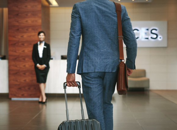 Photo Of A Man In A Dark Grey Suit Wheeling His Suitcase Towards A Hotel Receptionist And Her Desk