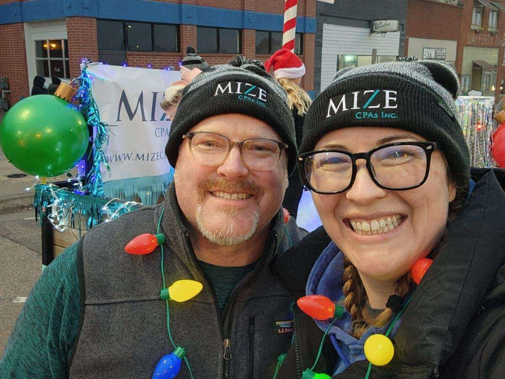 Two Mize Employees Wearing Mize Stocking Caps And A Necklace Of Holiday Lights Smiling In Front Of The North Pole Float